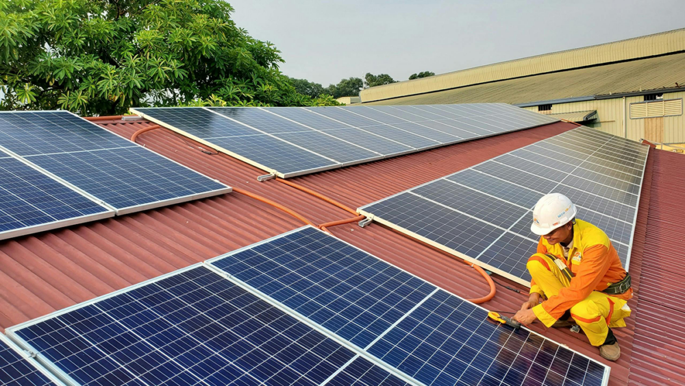 A worker in a yellow uniform and white helmet is installing solar panels on a red corrugated rooftop. Trees and a building are visible in the background.